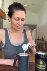 A woman in a gray tank top is shown in a home kitchen environment, holding a porcelain scoop over a dark speckled mug while adding Tocos / Vitamin E to a milk-like liquid. A Miron glass jar of Schizandu Tocos' is visible on the table next to her cup.
