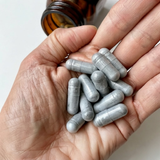 A close-up of a hand holding several gray oval supplement capsules, with part of an amber glass bottle visible at the top against a plain light background.