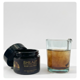 An open black jar labeled 'SHILAJIT RAW Live Resin' sits beside a clear glass beaker filled with a brown resin mixture on a light gray background.