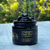 A jar of Wildcrafted Moor Mud Mask with a spoon resting on top, indicating the product is ready for use. The jar is labeled with information about the product being additive-free and sourced from the heart of Europe.