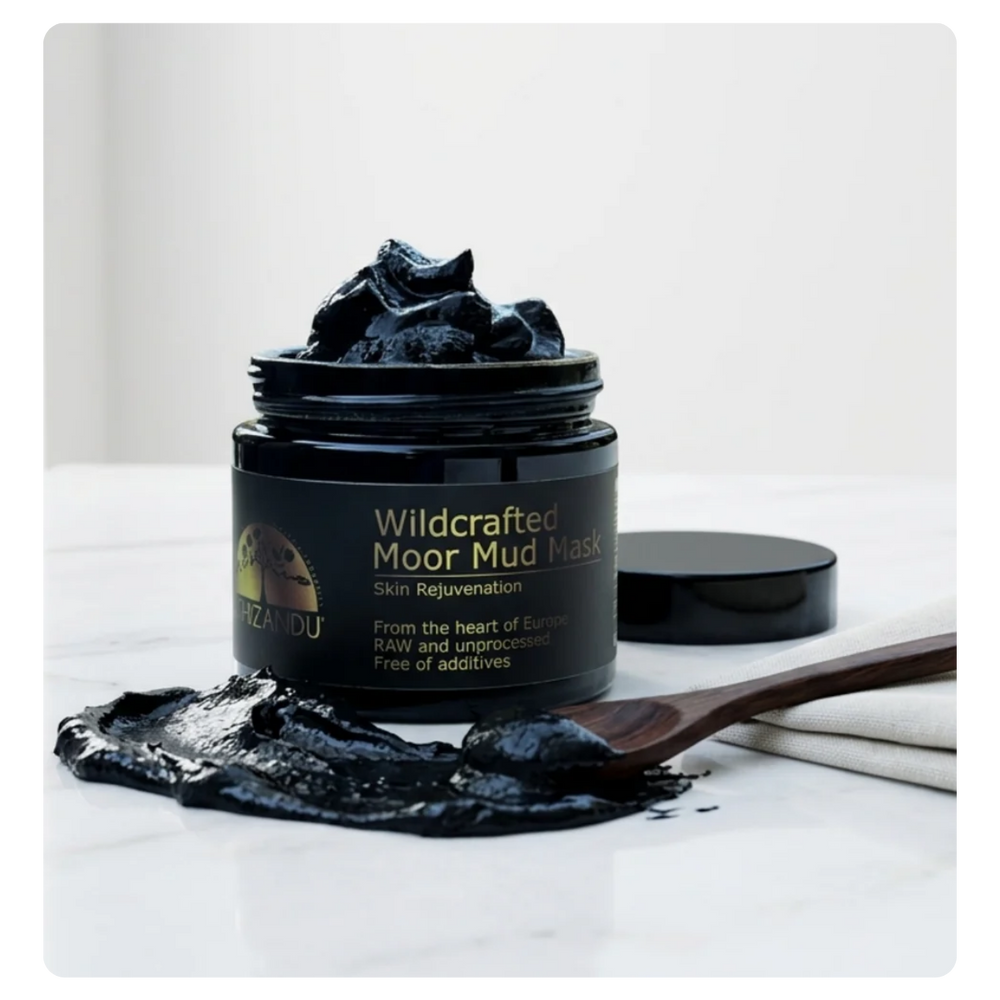 A jar of Wildcrafted Moor Mud Mask with black mud mask piled on top and on a wooden spoon, set on a white marble surface with the product lid beside it.