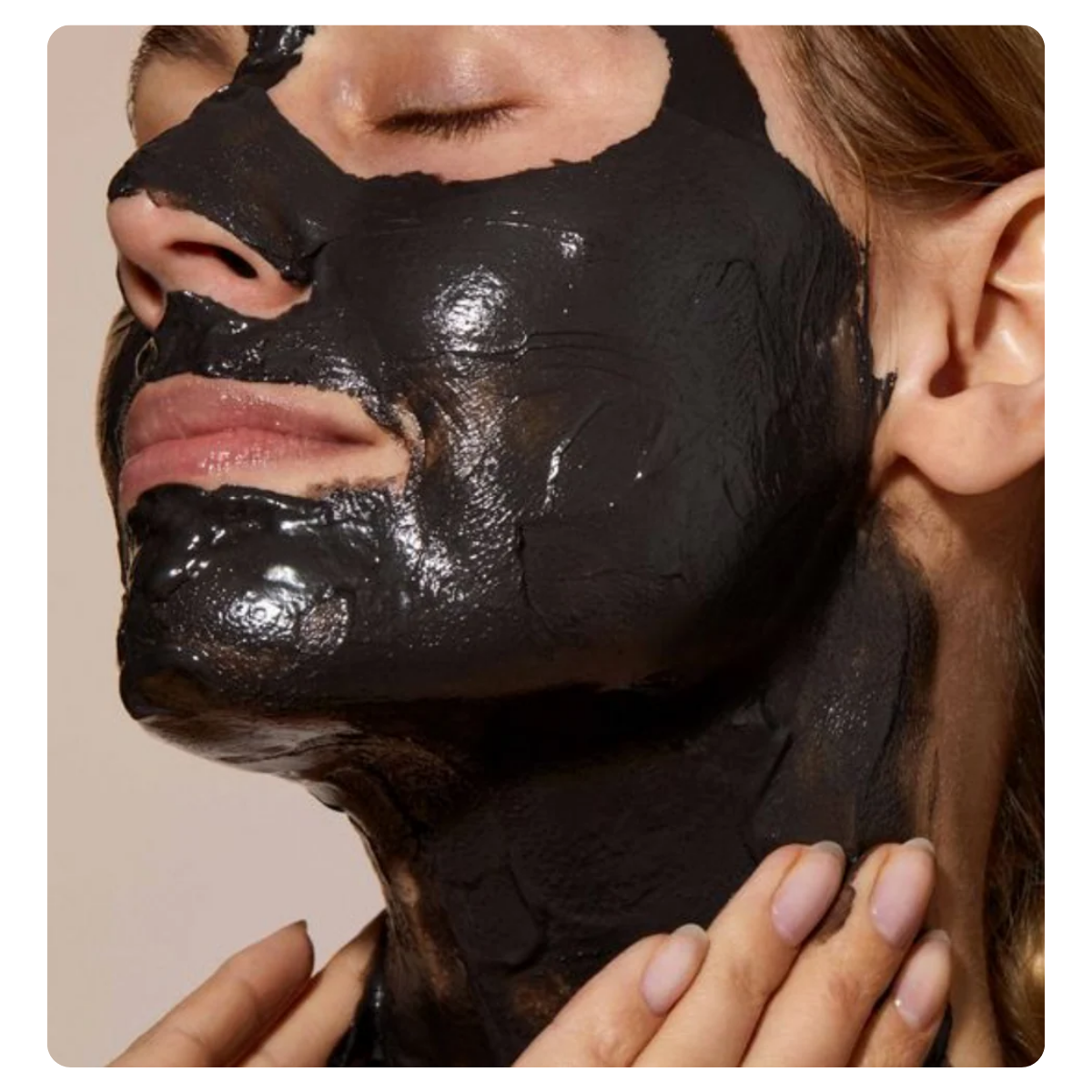 Close-up of a person wearing a thick, glossy black face mask, with hands touching the neck area against a plain beige background.