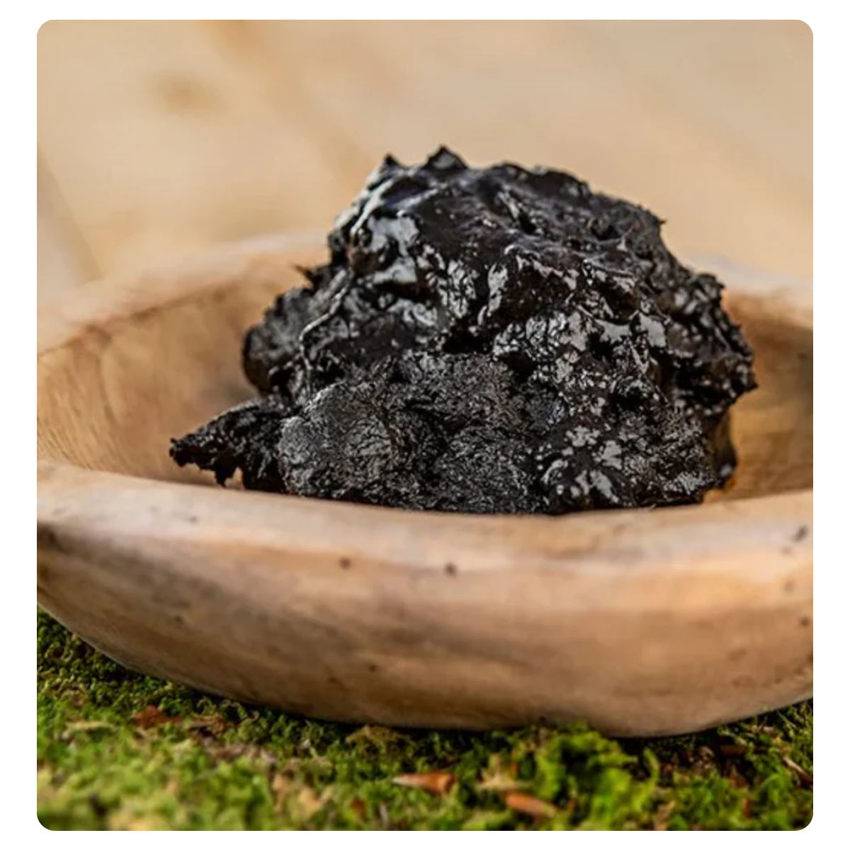 A close-up of a dark, crumbly textured food product, likely a nut butter or paste, resting in a light-colored wooden bowl on a surface with green moss or grass visible at the bottom.
