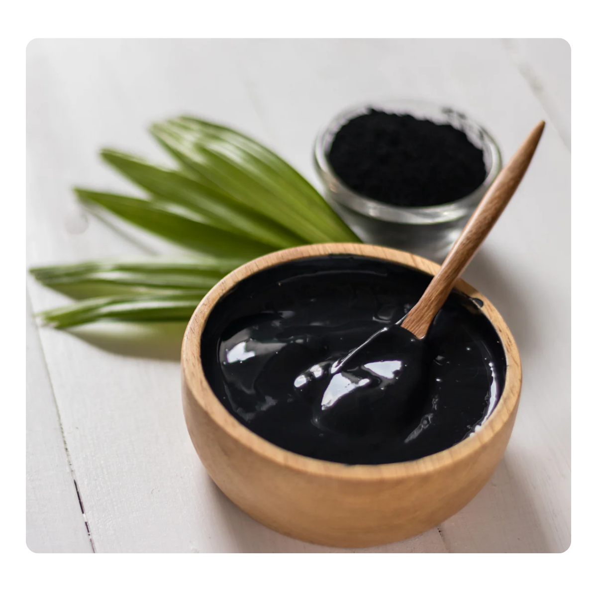 Black paste in a wooden bowl with a spoon, surrounded by green leaves on a white background