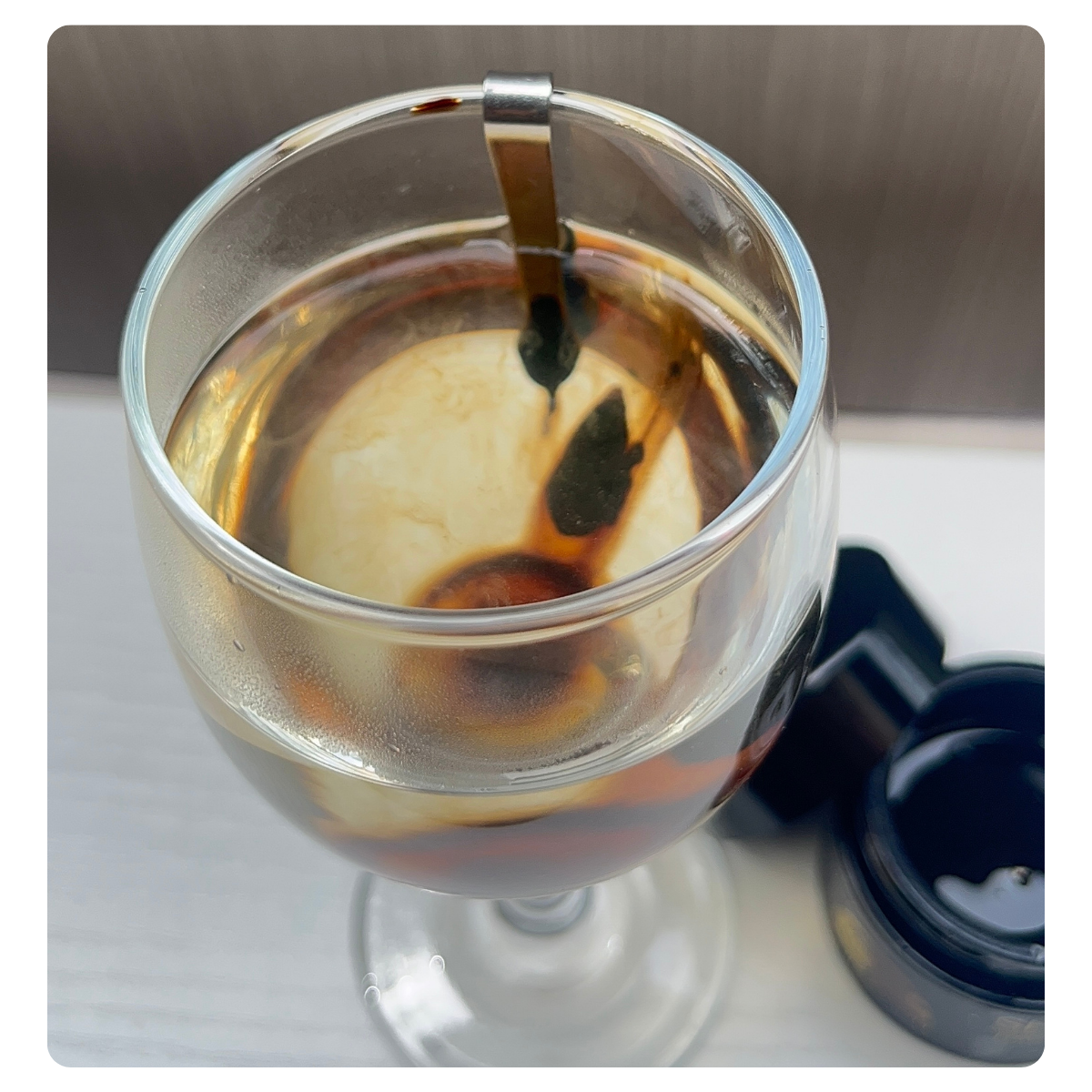 A clear glass cup containing a dark brown liquid, with a small metal spoon resting inside the cup. A black lid or container is visible at the lower right on a light surface, with a neutral gray background behind.