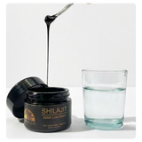 A jar of Shilajit RAW Live Resin with its lid off, showing a spoon dripping thick black resin above a glass of water, on a white background.