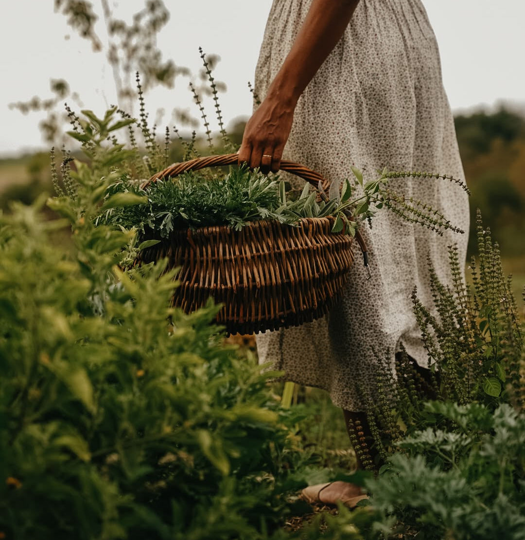 A woman in a light beige patterned dress stands in a lush green field, holding a woven wicker basket filled with leafy greens.
