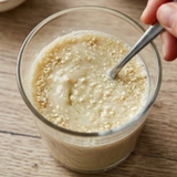 A close-up of Tocos / Vitamin E in a clear glass cup with a spoon, resting on a wooden surface. A hand is visible holding the spoon, stirring the Tocos into the milk-like liquid.
